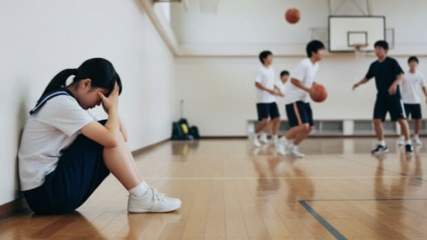 体育の授業についていけず、体育館の隅で一人座り込む起立性調節障害の生徒。