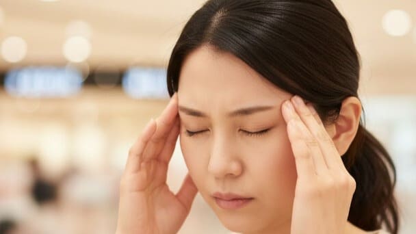 
両こめかみを指で押さえ、拍動性の頭痛に耐える日本人女性